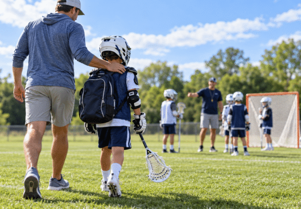 Child carrying a Lacrosse stick walks beside a parent toward their first Lacrosse practice on a sunny field.